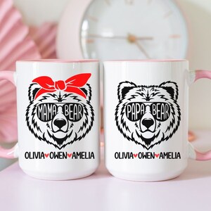 Op de afbeelding: Twee witte keramische mokken met roze randen. De mok aan de linkerkant heeft een zwart-witte illustratie van een beer met een zonnebril en een rode bandana met de tekst "Mama Bear" en de namen "Olivia Owen Amelia" eronder. De mok aan de rechterkant heeft een zwart-witte illustratie van een beer met een zonnebril met de tekst "Papa Bear" en de namen "Olivia Owen Amelia" eronder.