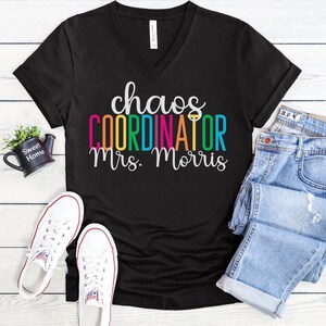 Pu&ograve; includere: Maglietta nera a collo a V con il testo "chaos coordinator mrs. morris" in colori arcobaleno.