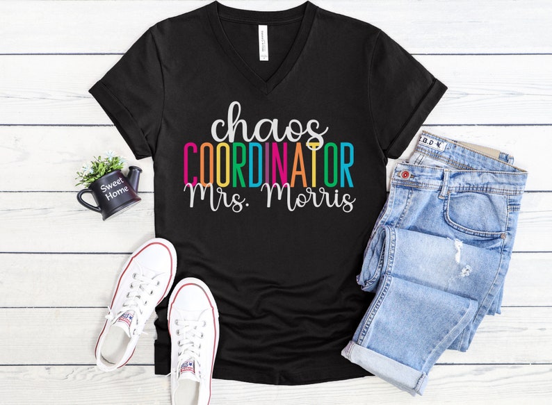 Pu&ograve; includere: Maglietta nera a collo a V con il testo "chaos coordinator mrs. morris" in colori arcobaleno.
