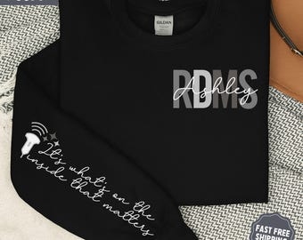 Può includere: Felpa nera con la scritta "RDMS Ashley" in bianco e grigio. La manica sinistra presenta una grafica a ultrasuoni e la frase "It's what's on the inside that matters" in corsivo bianco. La felpa è etichettata "Super Soft".