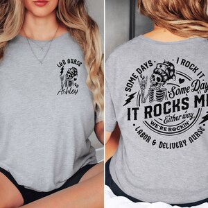 K&ouml;nnte beinhalten: Graues T-Shirt mit einem Skelett-Motiv und dem Text "LED NURSE Ashley" auf der Vorderseite und "SOME DAYS I ROCK IT Some Days IT ROCKS ME Either way WE'RE ROCKIN' LABOR & DELIVERY NURSE" auf der R&uuml;ckseite.