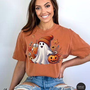 May include: A burnt orange t-shirt with a white ghost wearing a brown and orange knit hat holding a glass of orange juice. The ghost is surrounded by fall leaves and a jack-o-lantern.