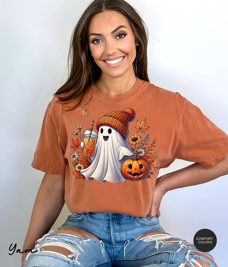 May include: A burnt orange t-shirt with a white ghost wearing a brown and orange knit hat holding a glass of orange juice. The ghost is surrounded by fall leaves and a jack-o-lantern.