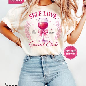 May include: A white t-shirt with the text "Self Love Social Club" in pink lettering. The design features a pink wine glass with a pink bow and glitter accents. The text "Est. 2025" is also included in the design.