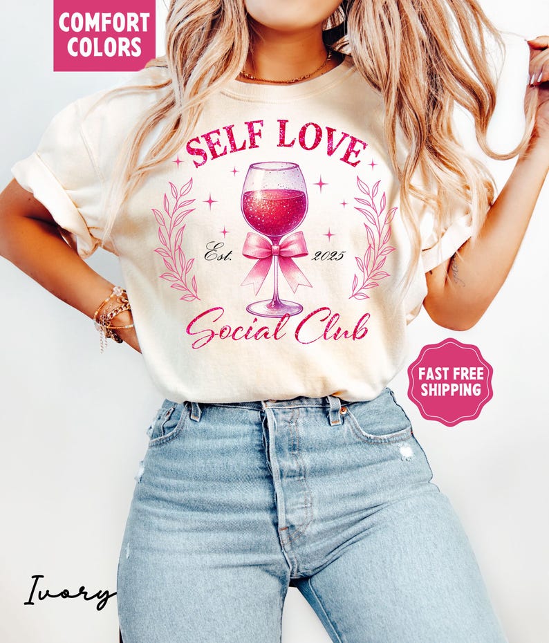 May include: A white t-shirt with the text "Self Love Social Club" in pink lettering. The design features a pink wine glass with a pink bow and glitter accents. The text "Est. 2025" is also included in the design.