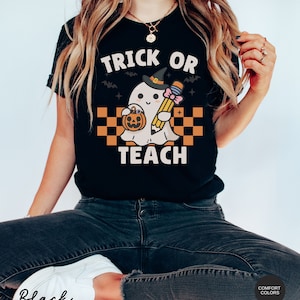 K&ouml;nnte beinhalten: Schwarzes T-Shirt mit einem Grafikdruck eines Geistes, der einen Hexenhut tr&auml;gt und einen Bleistift und einen K&uuml;rbis h&auml;lt. Der Text "Trick or Teach" ist auf dem T-Shirt aufgedruckt.