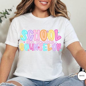 May include: White t-shirt with the words "SCHOOL Mrs. Kennedy COUNSELOR" in colorful bubble letters. The design includes flowers and stars. The shirt is a short-sleeved crew neck.