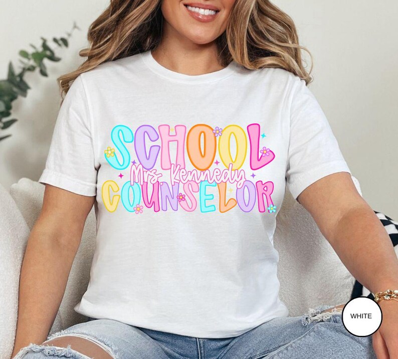 May include: White t-shirt with the words "SCHOOL Mrs. Kennedy COUNSELOR" in colorful bubble letters. The design includes flowers and stars. The shirt is a short-sleeved crew neck.