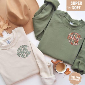 May include: Two cream colored sweatshirts with a floral monogram design. The monogram is in a circle and features a floral pattern. The sweatshirts are on a white background with a cup of coffee and fall leaves.