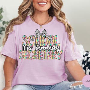Może przedstawiać: Fioletowy t-shirt z kolorowym wzorem "Sekretarka szkolna" z kokardką w czarne i białe kropki. Wz&oacute;r zawiera tekst "Pani Kennedy".