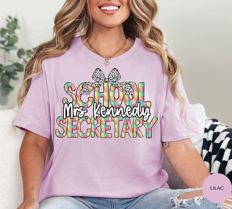 Może przedstawiać: Fioletowy t-shirt z kolorowym wzorem "Sekretarka szkolna" z kokardką w czarne i białe kropki. Wz&oacute;r zawiera tekst "Pani Kennedy".