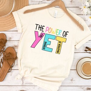 May include: Cream-coloured t-shirt with the text "THE POWER OF YET" in black and colourful block letters. The shirt is displayed on a wooden hanger. Sandals, a hat, and a cup of coffee are also visible.