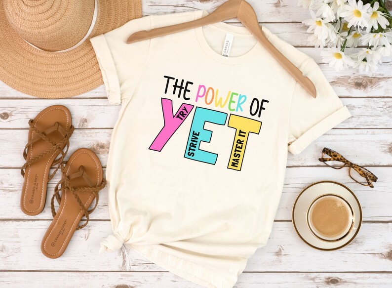 May include: Cream-coloured t-shirt with the text "THE POWER OF YET" in black and colourful block letters. The shirt is displayed on a wooden hanger. Sandals, a hat, and a cup of coffee are also visible.