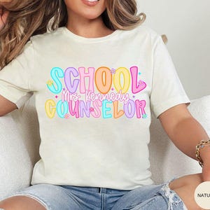 May include: Cream-colored t-shirt with the words "SCHOOL Mrs. Kennedy COUNSELOR" in colorful bubble letters. The text is decorated with small flowers and stars. The shirt is made of a soft material.