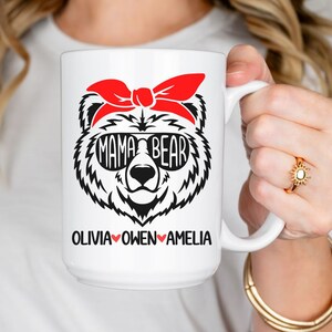 Op de afbeelding: Een witte keramische mok met een zwart-witte illustratie van een beer die een rode bandana en een zonnebril draagt. De tekst "Mama Bear" staat op de zonnebril. De tekst "Olivia Owen Amelia" staat onder de beer met een rood hart tussen elke naam.