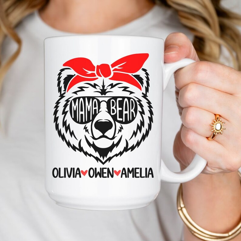 Op de afbeelding: Een witte keramische mok met een zwart-witte illustratie van een beer die een rode bandana en een zonnebril draagt. De tekst "Mama Bear" staat op de zonnebril. De tekst "Olivia Owen Amelia" staat onder de beer met een rood hart tussen elke naam.