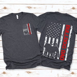 May include: Two dark heather grey t-shirts with a distressed American flag design. One shirt has a bulldog silhouette and the text "Bulldog Dad" in red. The other shirt has a smaller distressed American flag with a bulldog silhouette.