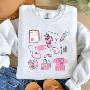 K&ouml;nnte beinhalten: Ein hellgraues Sweatshirt mit rosa medizinischen Illustrationen. Das Design umfasst ein Stethoskop, eine Gesichtsmaske, eine Krankenschwesterm&uuml;tze, einen Klemmbrett, medizinische Werkzeuge und eine rosa Wasserflasche. Das Sweatshirt wird mit blauen Jeans und Sandalen kombiniert.