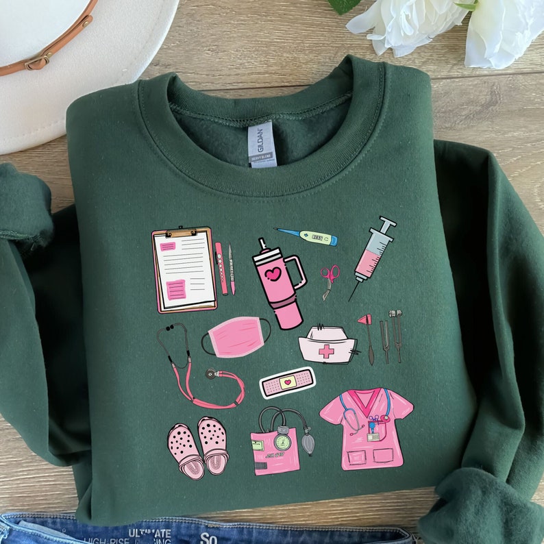 K&ouml;nnte beinhalten: Dunkelgr&uuml;nes Sweatshirt mit einer Sammlung von rosa und wei&szlig;en medizinischen Illustrationen. Das Design umfasst ein Stethoskop, einen Klemmbrett, eine Gesichtsmaske, eine Krankenschwesterm&uuml;tze und eine Wasserflasche mit einem Herz. Das Sweatshirt ist auf einer Holzoberfl&auml;che gefaltet.