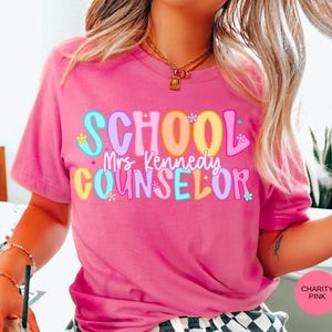 May include: A pink t-shirt with the words "SCHOOL Mrs. Kennedy COUNSELOR" in colorful, bubble letters. The shirt is a short-sleeved crew neck and is made of a soft material.