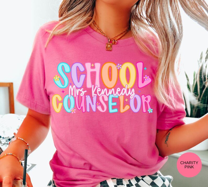 May include: A pink t-shirt with the words "SCHOOL Mrs. Kennedy COUNSELOR" in colorful, bubble letters. The shirt is a short-sleeved crew neck and is made of a soft material.