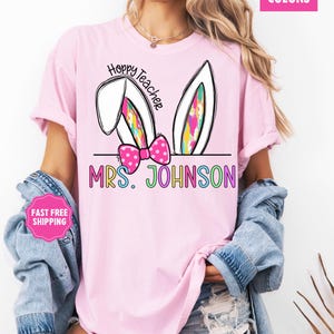 May include: Pink t-shirt with a graphic of bunny ears and a bow. The text "Hoppy Teacher" is above the bunny ears and "MRS. JOHNSON" is below the bunny ears. The text is in rainbow colors.