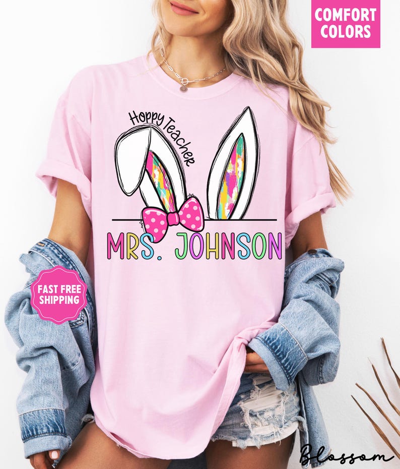 May include: Pink t-shirt with a graphic of bunny ears and a bow. The text "Hoppy Teacher" is above the bunny ears and "MRS. JOHNSON" is below the bunny ears. The text is in rainbow colors.