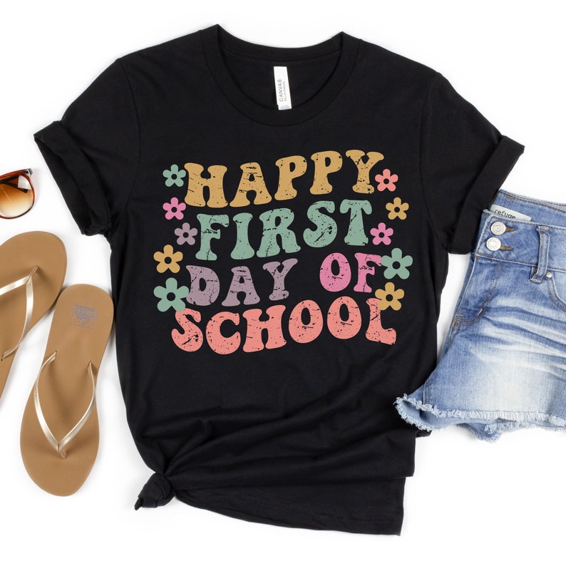 Puede incluir: Camiseta negra con el texto "Happy First Day of School" en fuente retro de colores con flores alrededor del texto.