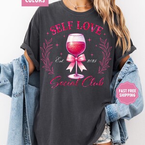 May include: A dark gray t-shirt with a pink glitter design that says "Self Love Social Club" with a wine glass and a pink bow. The design also includes the text "Est. 2023".
