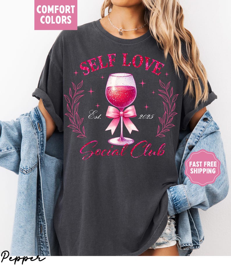 May include: A dark gray t-shirt with a pink glitter design that says "Self Love Social Club" with a wine glass and a pink bow. The design also includes the text "Est. 2023".