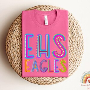 Puede incluir: Camiseta rosa con el texto "EHS EAGLES" en letras de colores y contorneadas. Las letras est&aacute;n decoradas con peque&ntilde;os puntos de colores.