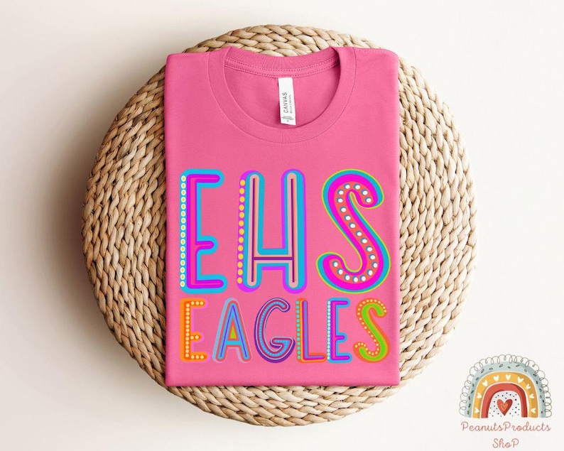 Puede incluir: Camiseta rosa con el texto "EHS EAGLES" en letras de colores y contorneadas. Las letras est&aacute;n decoradas con peque&ntilde;os puntos de colores.