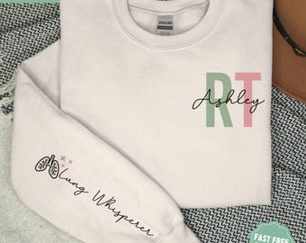 Può includere: Una felpa bianca con il testo "Ashley RT" sul davanti e "Lung Whisperer" sulla manica sinistra. Il testo è in nero e le lettere "RT" sono in verde e rosa.