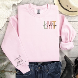 May include: Pink sweatshirt with the text "Ashley LMT" on the front and "muscle whisperer" on the left sleeve.