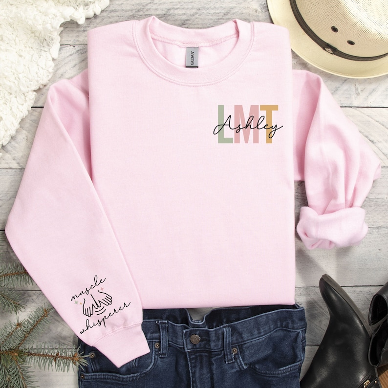 May include: Pink sweatshirt with the text "Ashley LMT" on the front and "muscle whisperer" on the left sleeve.