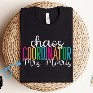 Pu&ograve; includere: Maglietta nera con il testo "chaos coordinator Mrs. Morris" in colori arcobaleno. Il testo &egrave; su una maglietta nera. La maglietta &egrave; piegata e mostrata su un cesto intrecciato.