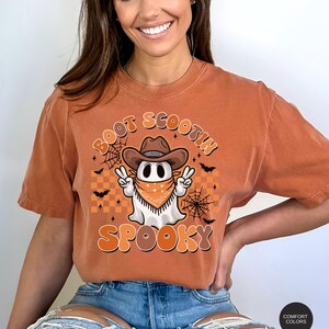 May include: A burnt orange t-shirt with a cartoon ghost wearing a cowboy hat and bandana. The ghost is holding up two peace signs. The text "Boot Scootin' Spooky" is printed on the shirt.
