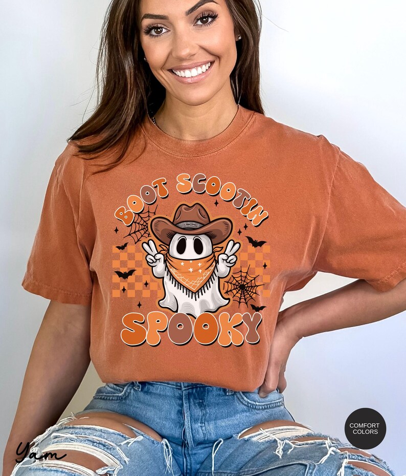 May include: A burnt orange t-shirt with a cartoon ghost wearing a cowboy hat and bandana. The ghost is holding up two peace signs. The text "Boot Scootin' Spooky" is printed on the shirt.