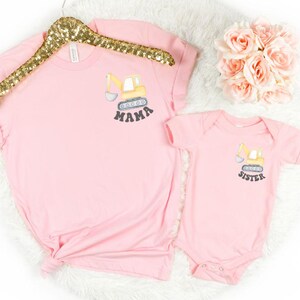 Peut inclure: Ensemble rose compos&eacute; d'un t-shirt et d'un body pour b&eacute;b&eacute;. Le t-shirt porte le mot "MAMA" et le body le mot "SISTER" imprim&eacute;s sous un dessin de v&eacute;hicule de chantier jaune. L'ensemble est pr&eacute;sent&eacute; sur une surface blanche avec un cintre dor&eacute; &agrave; paillettes.
