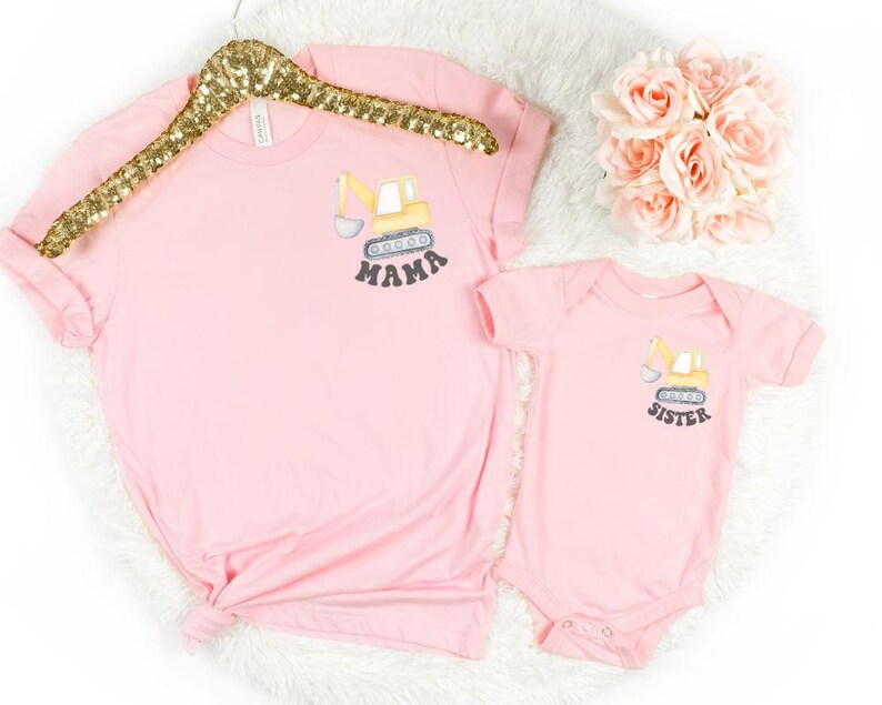 Peut inclure: Ensemble rose compos&eacute; d'un t-shirt et d'un body pour b&eacute;b&eacute;. Le t-shirt porte le mot "MAMA" et le body le mot "SISTER" imprim&eacute;s sous un dessin de v&eacute;hicule de chantier jaune. L'ensemble est pr&eacute;sent&eacute; sur une surface blanche avec un cintre dor&eacute; &agrave; paillettes.