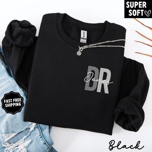 May include: Black crewneck sweatshirt with the text "DR Jones" in gray and white. A silver necklace with a circular pendant rests on the sweatshirt. The words "Super Soft" and "Black" are also visible.