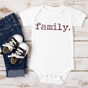 May include: White baby onesie with short sleeves and the word "family" printed in maroon on the front. The onesie is lying on a white wooden surface with a pair of black and white baby shoes and blue jeans.