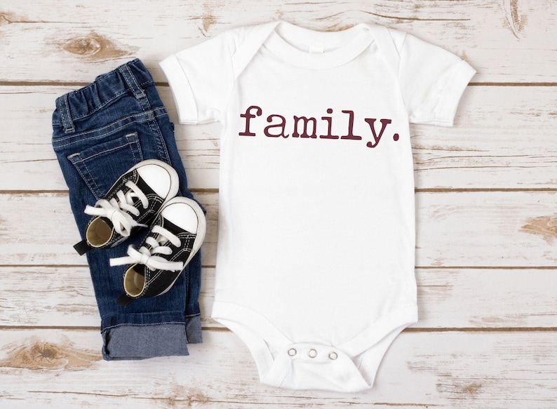 May include: White baby onesie with short sleeves and the word "family" printed in maroon on the front. The onesie is lying on a white wooden surface with a pair of black and white baby shoes and blue jeans.