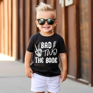 May include: A toddler boy wearing a black t-shirt with the text "Bad Two The Bone" in white. The shirt features a skeleton hand making a peace sign and a lightning bolt. The boy is wearing white shorts and black sunglasses.