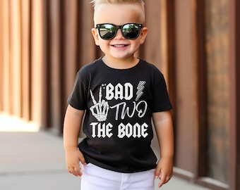 Peut inclure: Un t-shirt noir avec un motif blanc qui dit "Bad Two The Bone" avec une main de squelette faisant un signe de paix et un éclair. Un jeune garçon porte le t-shirt et des lunettes de soleil noires.