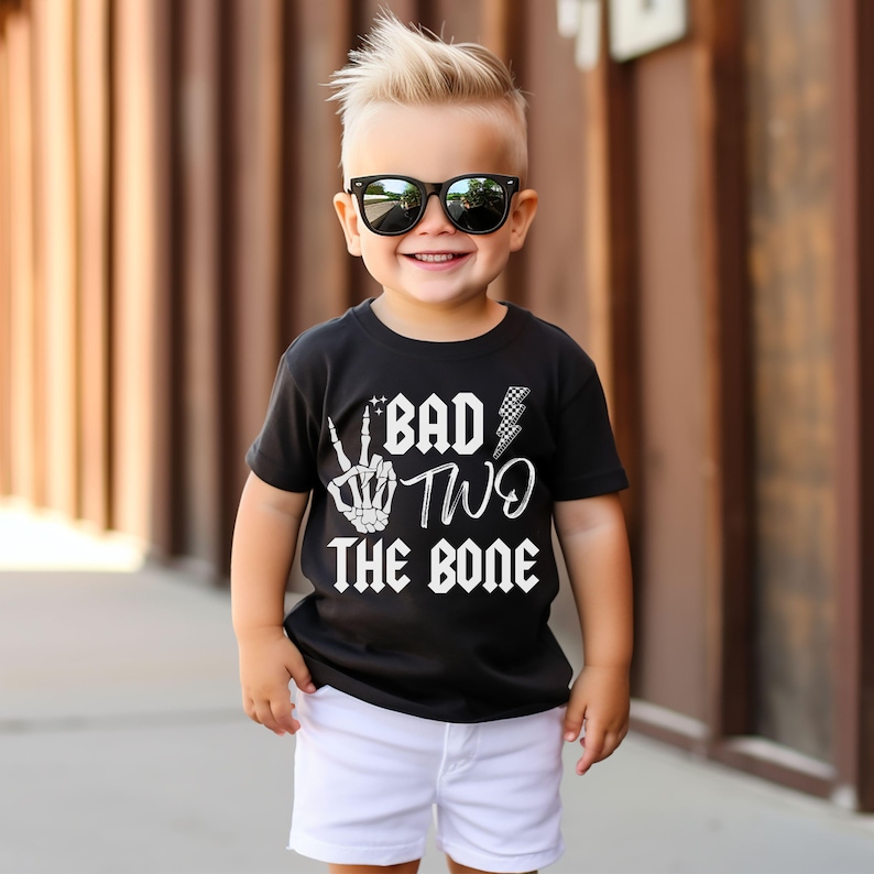 May include: A toddler boy wearing a black t-shirt with the text "Bad Two The Bone" in white. The shirt features a skeleton hand making a peace sign and a lightning bolt. The boy is wearing white shorts and black sunglasses.