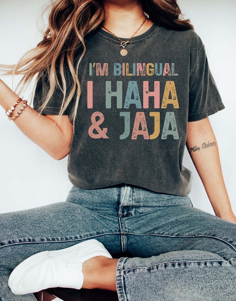 Puede incluir: Una camiseta gris oscuro con el texto "I'm Bilingual I HAHA & JAJA" impreso en letras coloridas y desgastadas.