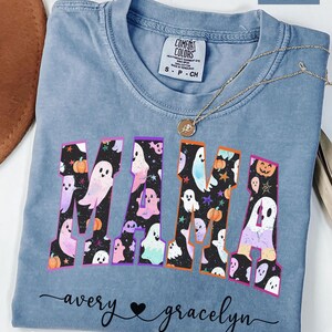 May include: Blue jean colored t-shirt with the word "MAMA" in a Halloween-themed design. The letters are filled with ghosts, pumpkins, and stars. The names "avery" and "gracelyn" are written below the word "MAMA".