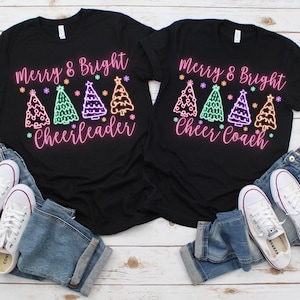 Pu&ograve; includere: Due magliette nere con il testo "Merry & Bright" e una fila di alberi di Natale colorati. Una maglietta dice "Cheerleader" e l'altra dice "Cheer Coach".