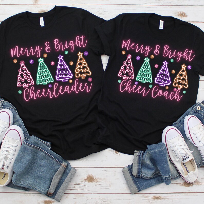 Pu&ograve; includere: Due magliette nere con il testo "Merry & Bright" e una fila di alberi di Natale colorati. Una maglietta dice "Cheerleader" e l'altra dice "Cheer Coach".
